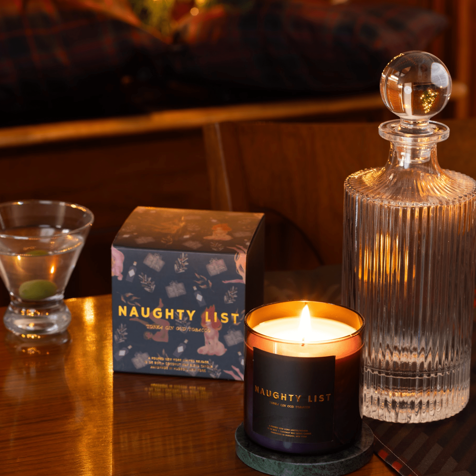 Candle with 'Naughty List' packaging on a table with a decanter and glass.