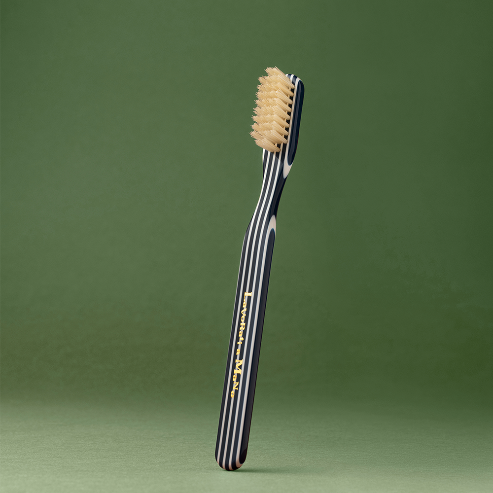 Blue Capri Collection Toothbrush with a dark navy handle featuring white stripes and gold lettering, standing upright against a solid green background.