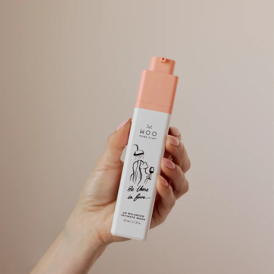 A hand holding a bottle of Woo More Play's ph Balanced Cleansing Wash against a solid beige background.