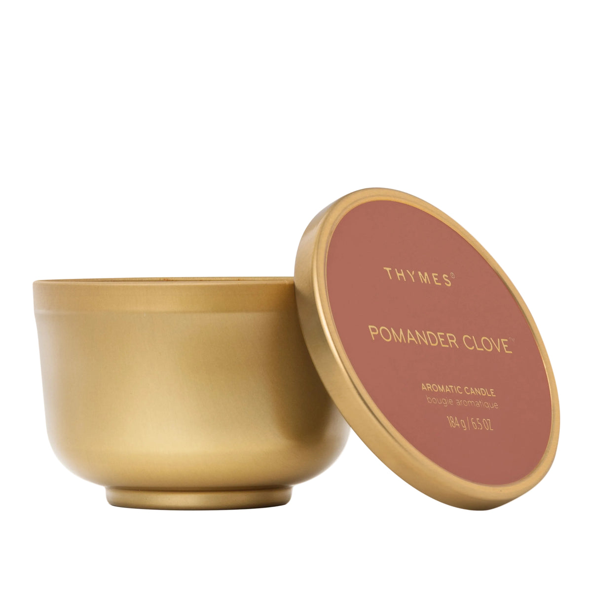 A gold metal candle jar with its lid leaning against it on a clean white background. The lid features a dusty rose label with gold text reading 'THYMES POMANDER CLOVE'.
