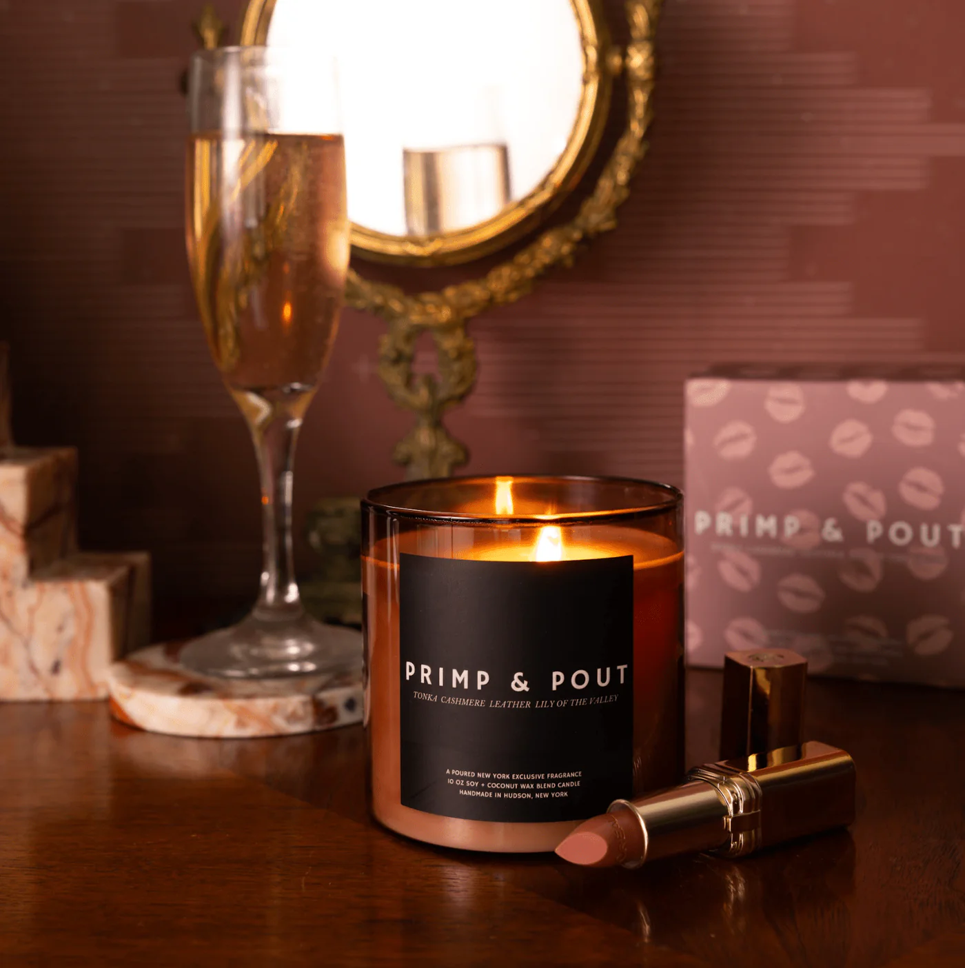 Candle with 'Primp & Pout' label on a table with a glass of champagne and makeup items.