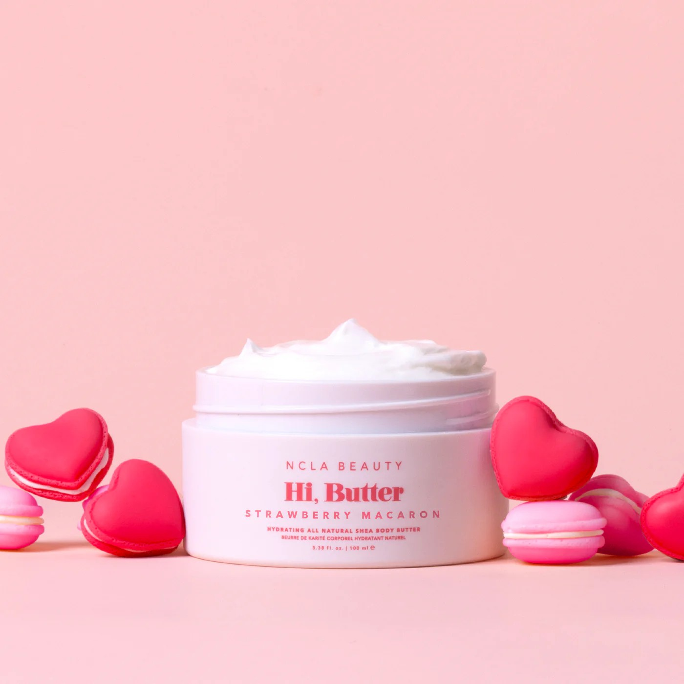 Jar of NCLA Beauty Hi, Butter Strawberry Macaron body butter with heart-shaped macarons on a pink background