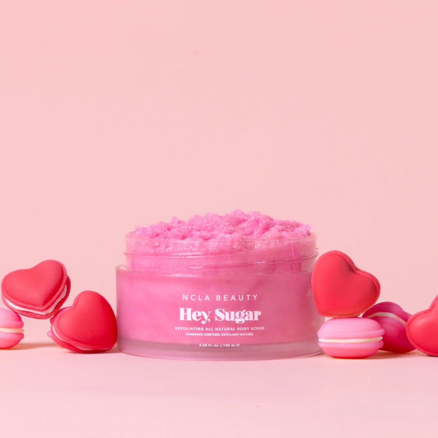 Pink body scrub jar with 'Hey Sugar' text on a pink background with heart-shaped macarons.