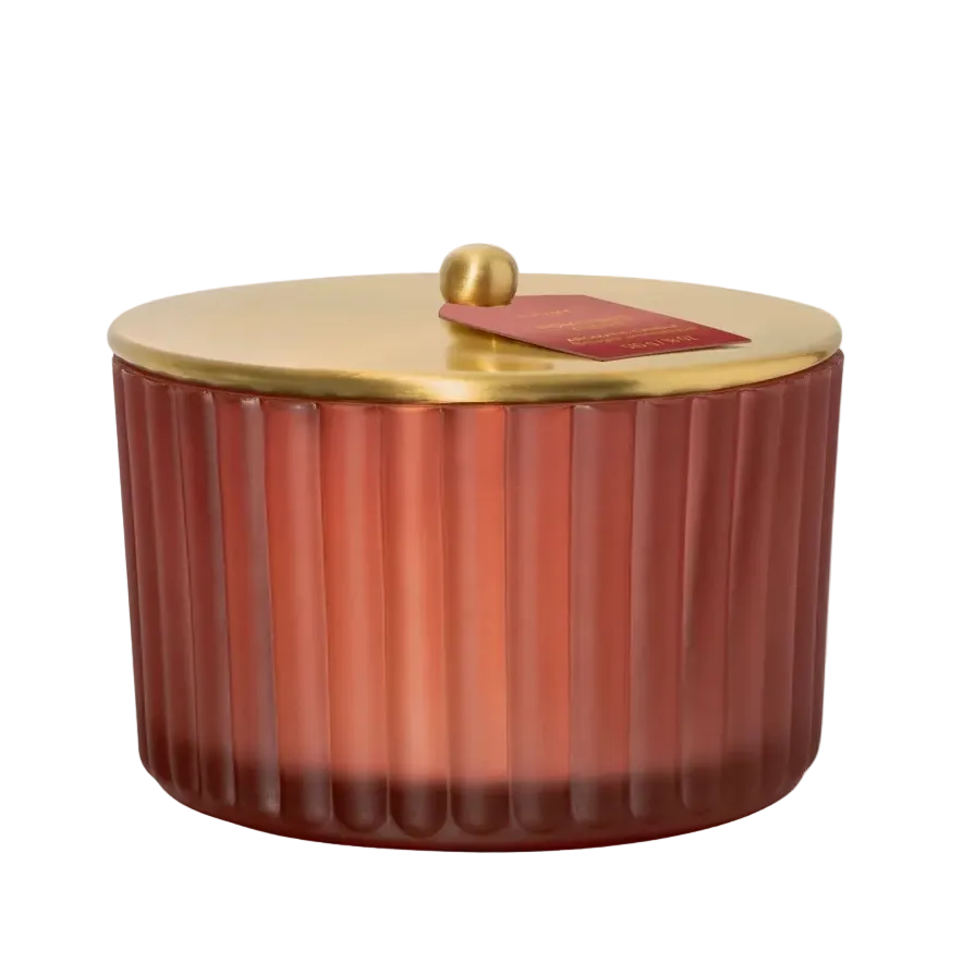 A ribbed glass candle jar in a warm reddish-brown hue with a polished gold metal lid and a small red wax tab, set against a clean white background.