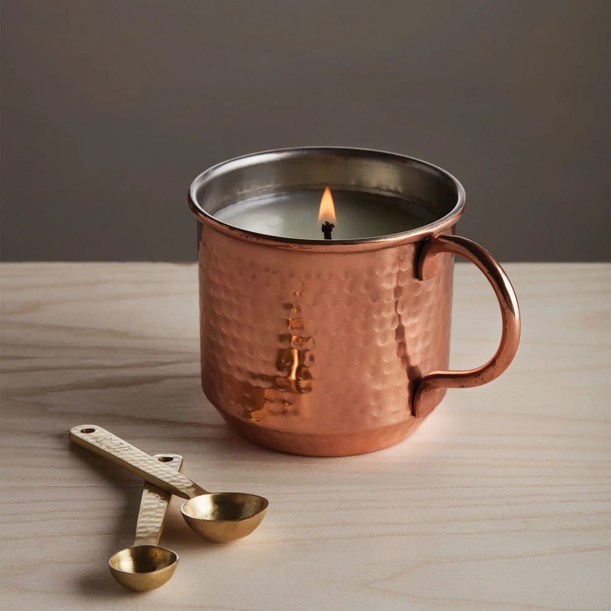 Copper mug with a lit candle inside on a wooden surface, accompanied by a brass measuring spoon.
