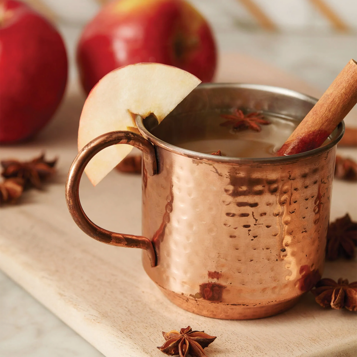 Copper mug with hot beverage, apple slice, and cinnamon stick on a wooden board with apples and star anise in the background.