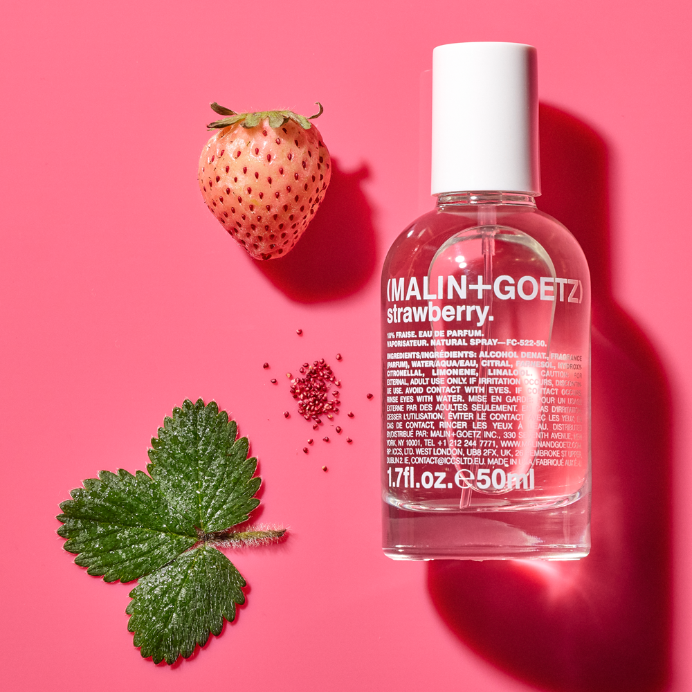 A clear glass bottle of MALIN+GOETZ strawberry Eau de Parfum with a white cap, styled on a vibrant pink background with a fresh strawberry, green leaves, and tiny red seeds.