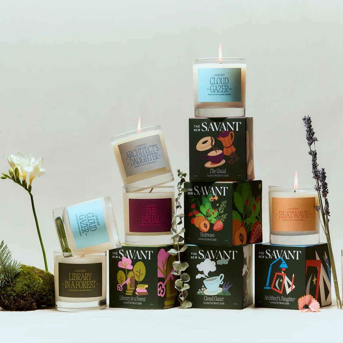 A styled product display of multiple Swant candles in glass jars and boxes, arranged in a pyramid with decorative moss, a white flower, and dried lavender on a light neutral background.