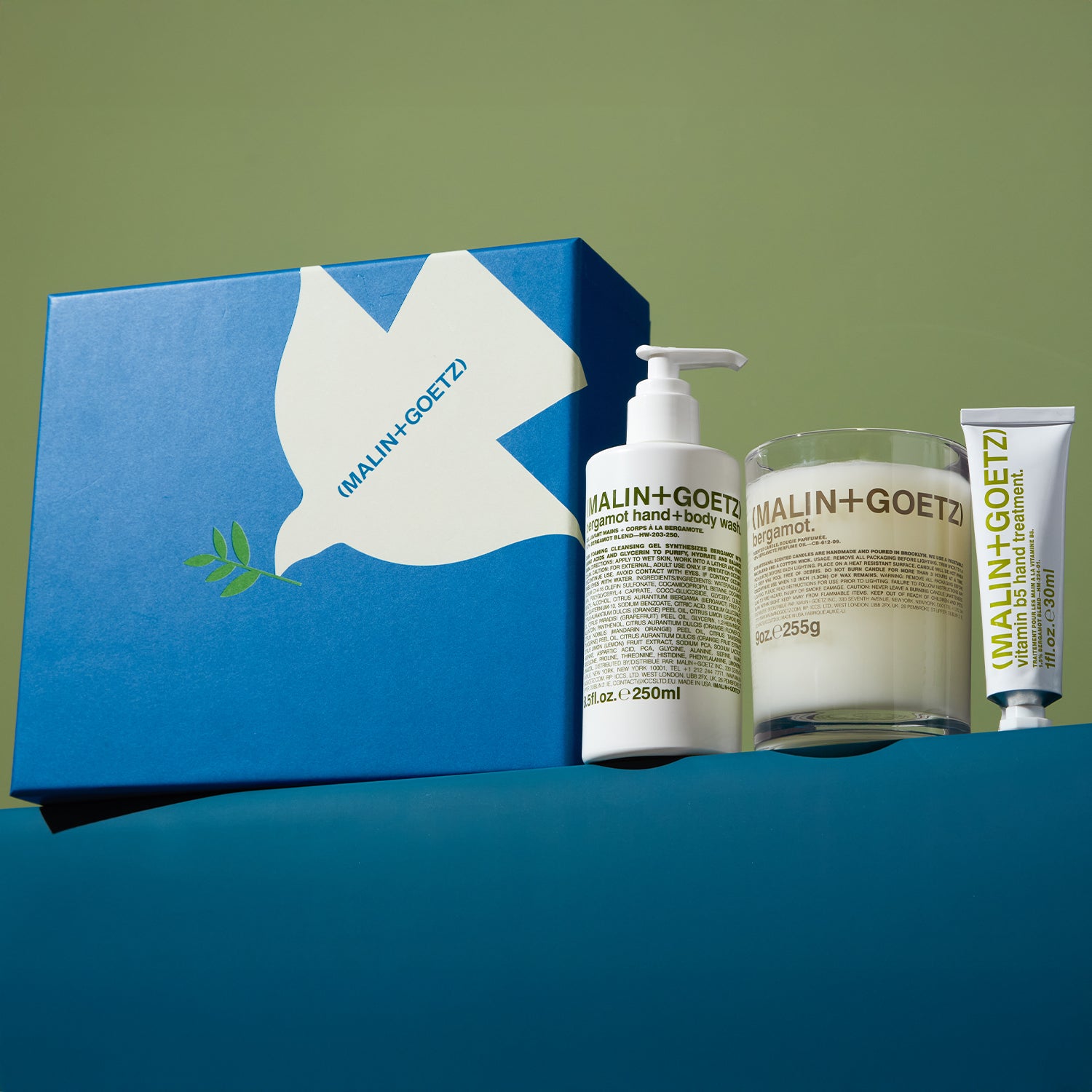 MALIN+GOETZ skincare products including a box, lotion, candle, and tube on a green and blue background.