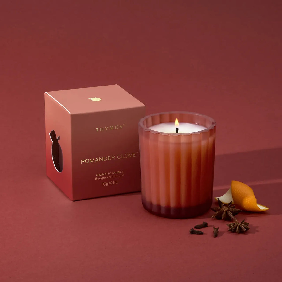 A lit Thymes Pomander Clove aromatic candle in a ribbed glass jar sits beside its matching pink box on a dusty rose background, with orange peel and star anise props nearby.