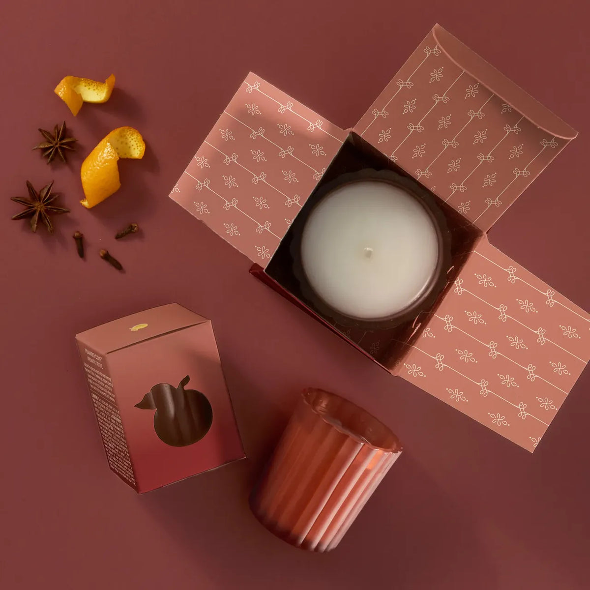 A candle product shown in a styled flat lay on a dusty rose background, with an open patterned box containing a white candle, a metallic rose-gold glass vessel, and small decorative spices and citrus peel.