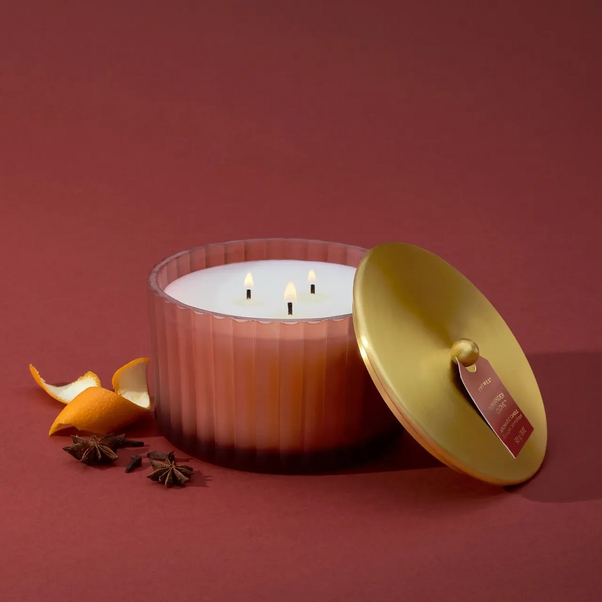 A three-wick candle in a ribbed clear glass jar with a gold-toned lid leaning beside it, styled on a dark red background with orange peel and star anise.
